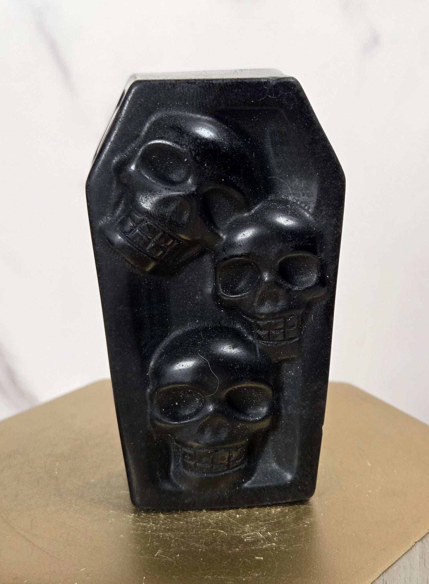 A black coffin-shaped decorative object with three carved skull motifs, displayed on a gold hexagonal pedestal against a light marble background.