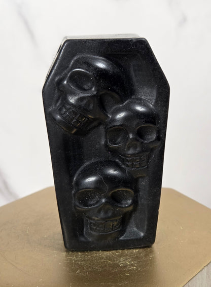 A black coffin-shaped decorative object with three carved skull motifs, displayed on a gold hexagonal pedestal against a light marble background.