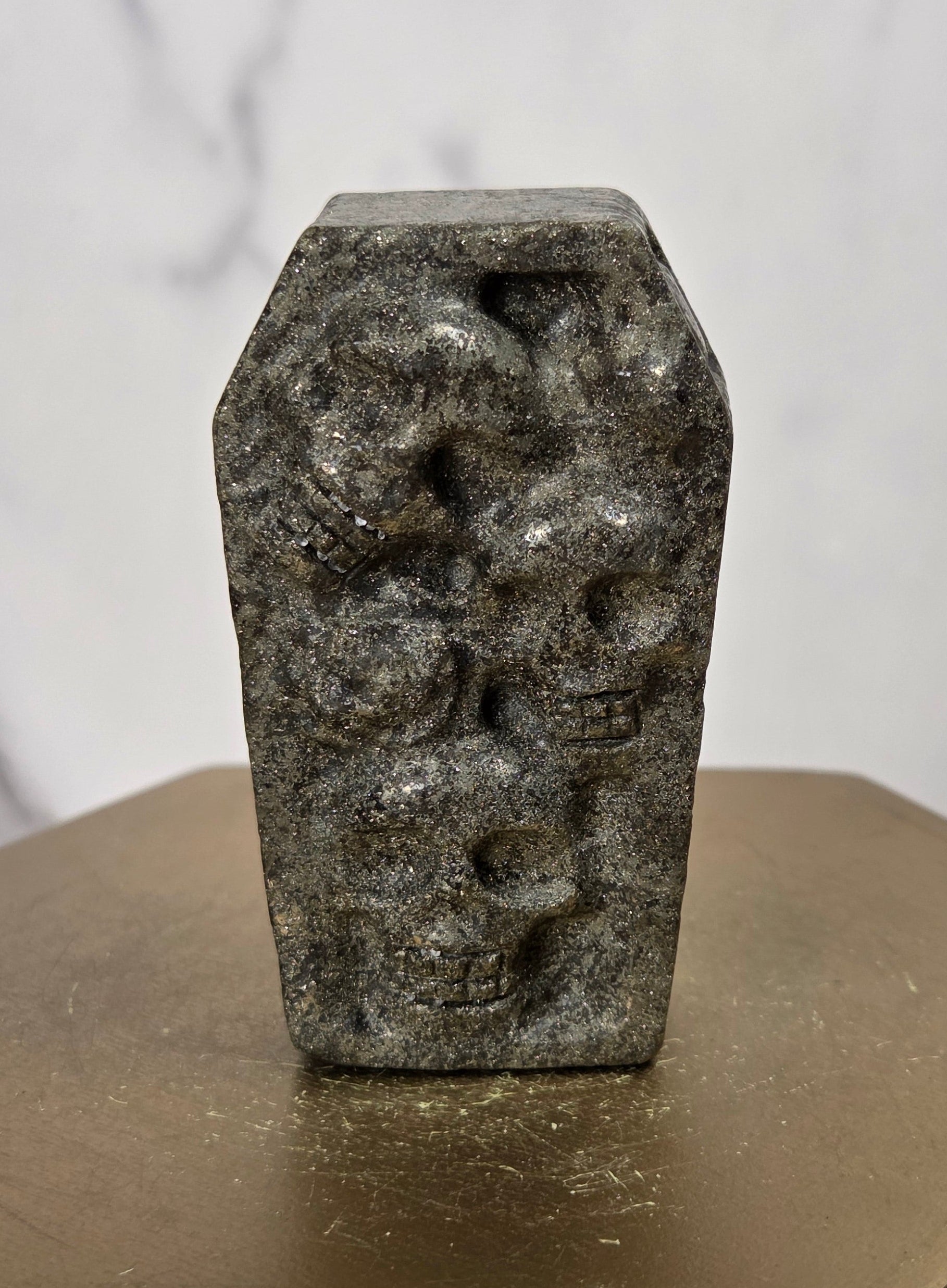 A small carved dark stone object with a coffin-shaped top and raised skull motifs, displayed upright on a metallic gold hexagonal pedestal against a light marble-patterned background.