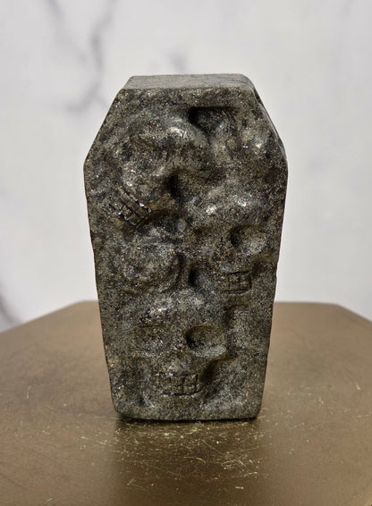 A small carved dark stone object with a coffin-shaped top and raised skull motifs, displayed upright on a metallic gold hexagonal pedestal against a light marble-patterned background.