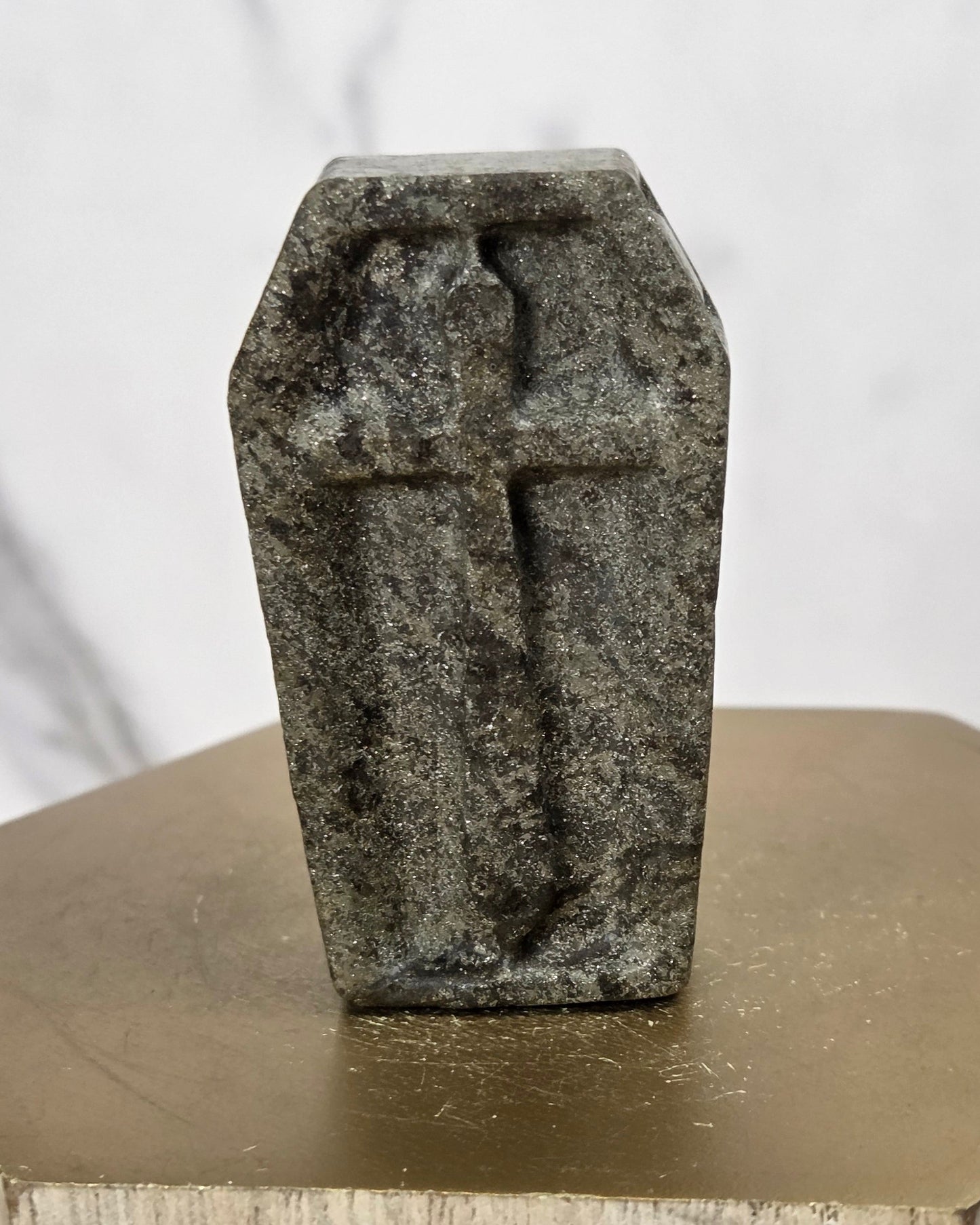 A small gray stone figurine shaped like a coffin with a cross on top, displayed on a hexagonal metallic gold pedestal against a light marble background.
