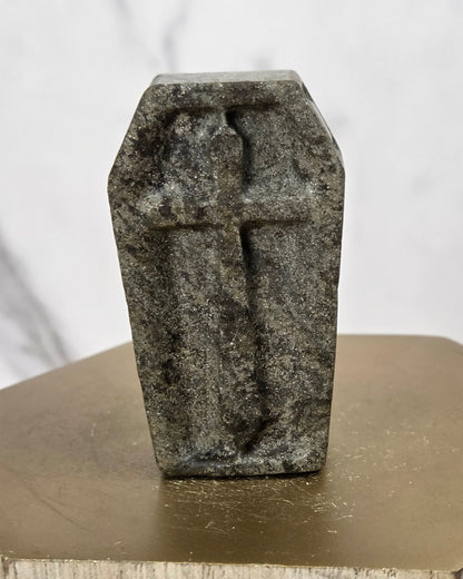 A small gray stone figurine shaped like a coffin with a cross on top, displayed on a hexagonal metallic gold pedestal against a light marble background.