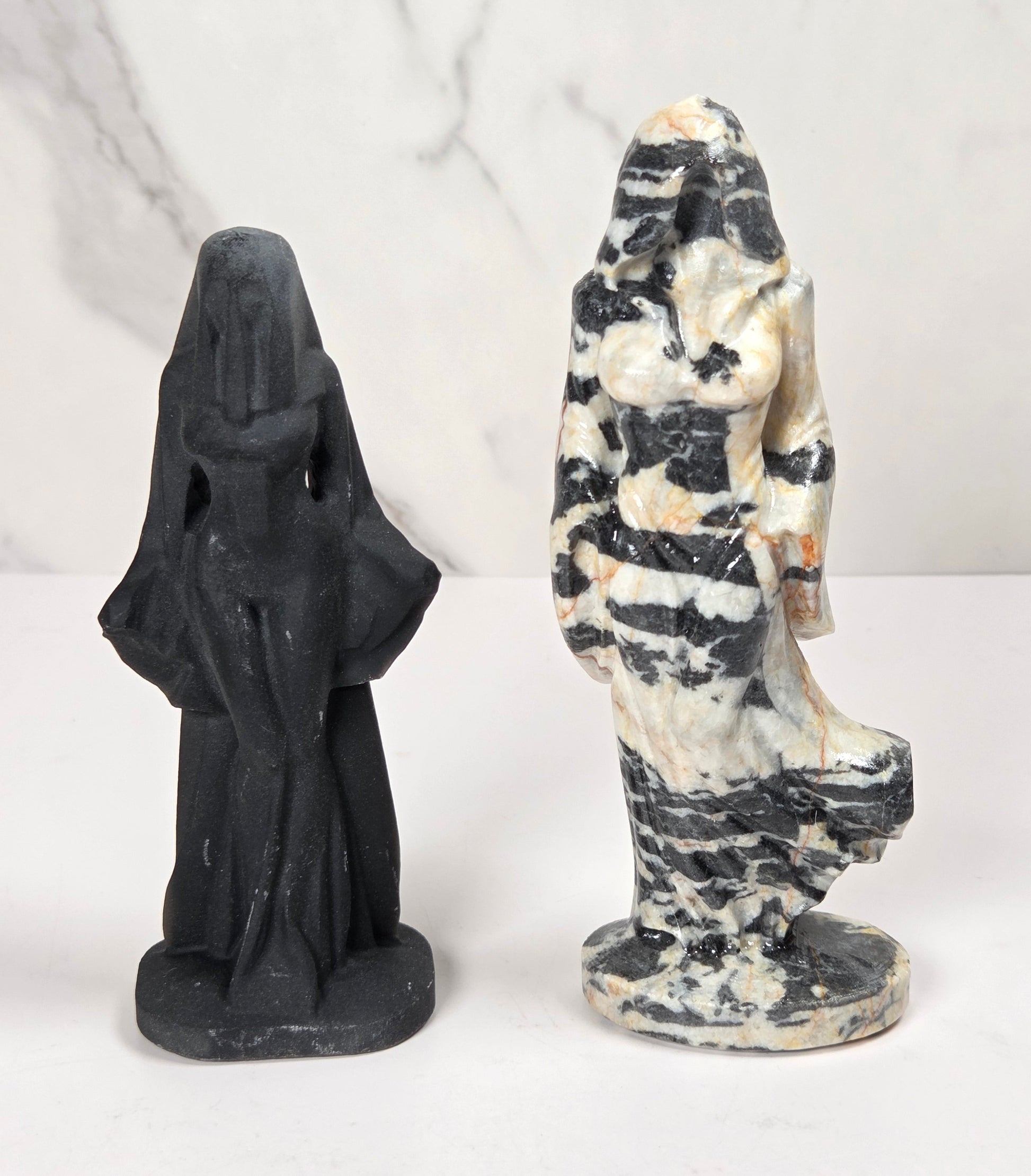 Two small carved stone figurines on a white surface against a white marble-look background. One figurine is matte black, and the other is a marbled black-and-white stone with beige areas.