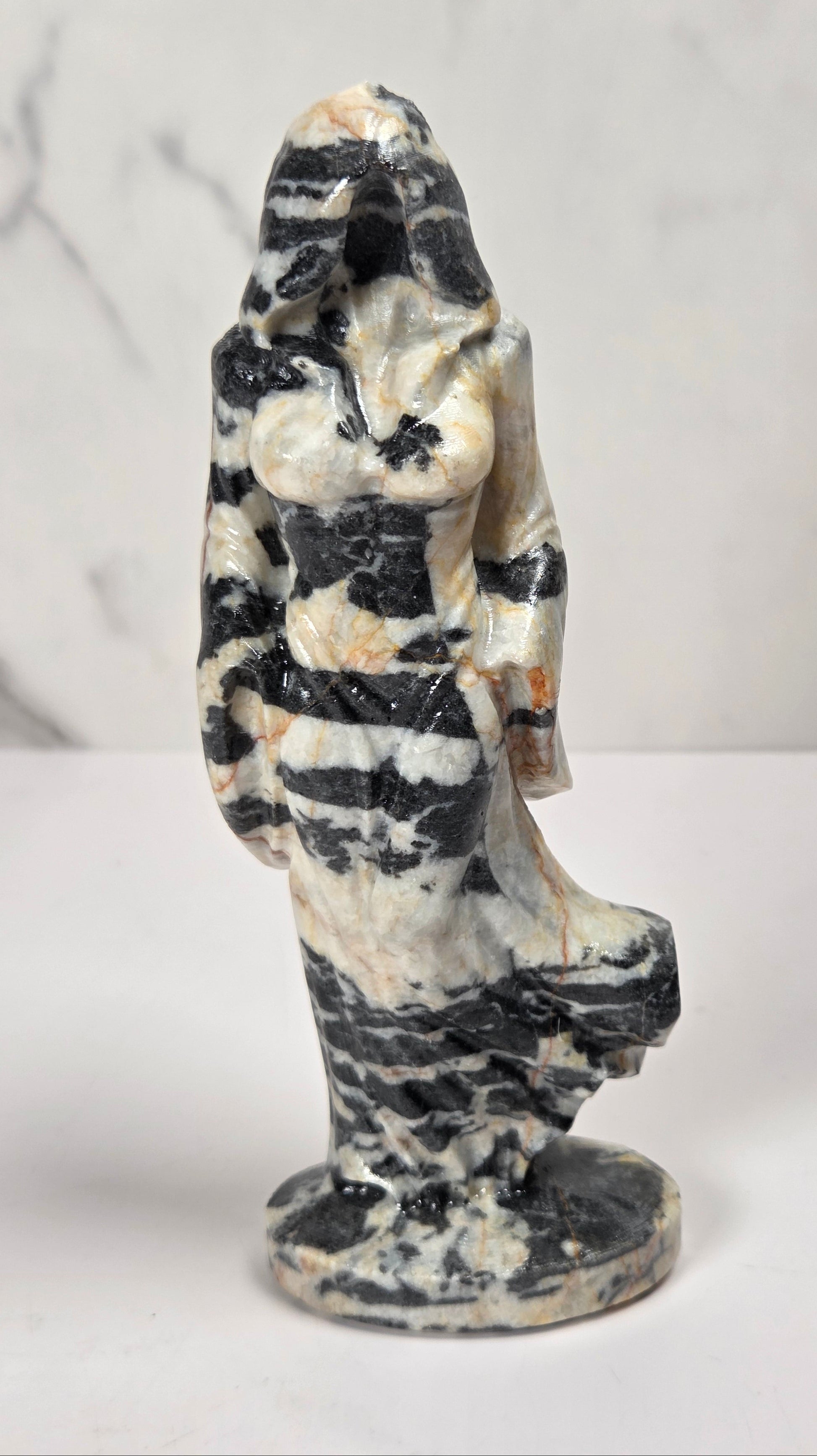 A small carved figurine of a hooded female figure standing on a round base, made from marbled stone with black, white, gray, and tan veining, photographed against a light marble-patterned background.