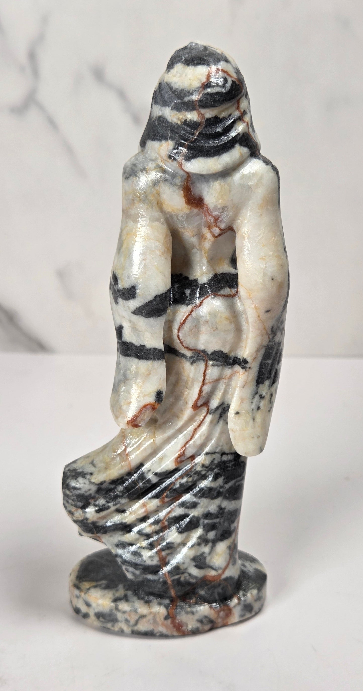 A small carved figurine of a robed female figure standing on a round base, made from marbled stone with cream, black, gray, and reddish-brown veining, photographed against a light marble-patterned background.