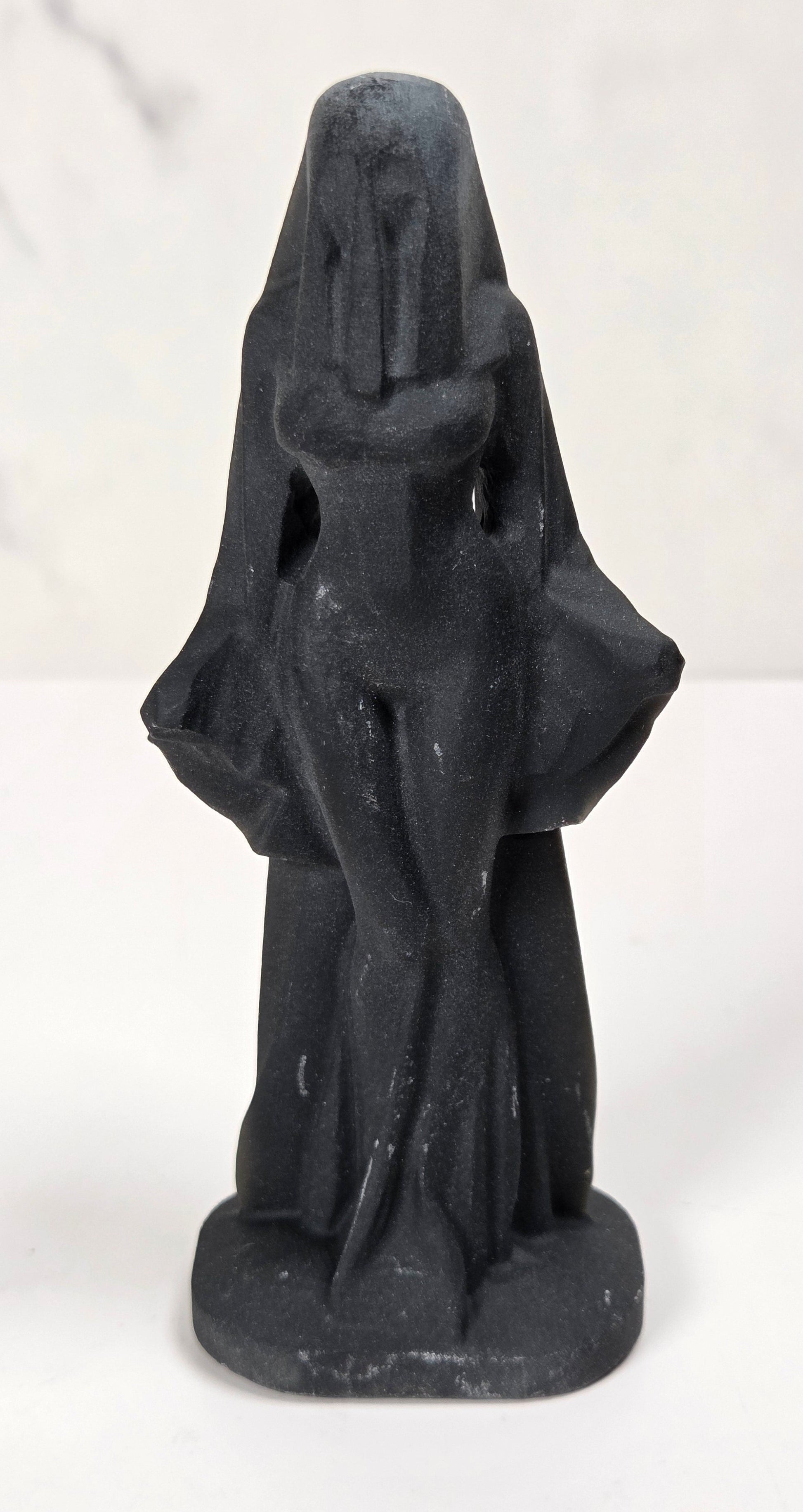 A small black figurine of a hooded, faceless figure standing on a round base, photographed against a light marble-patterned background.