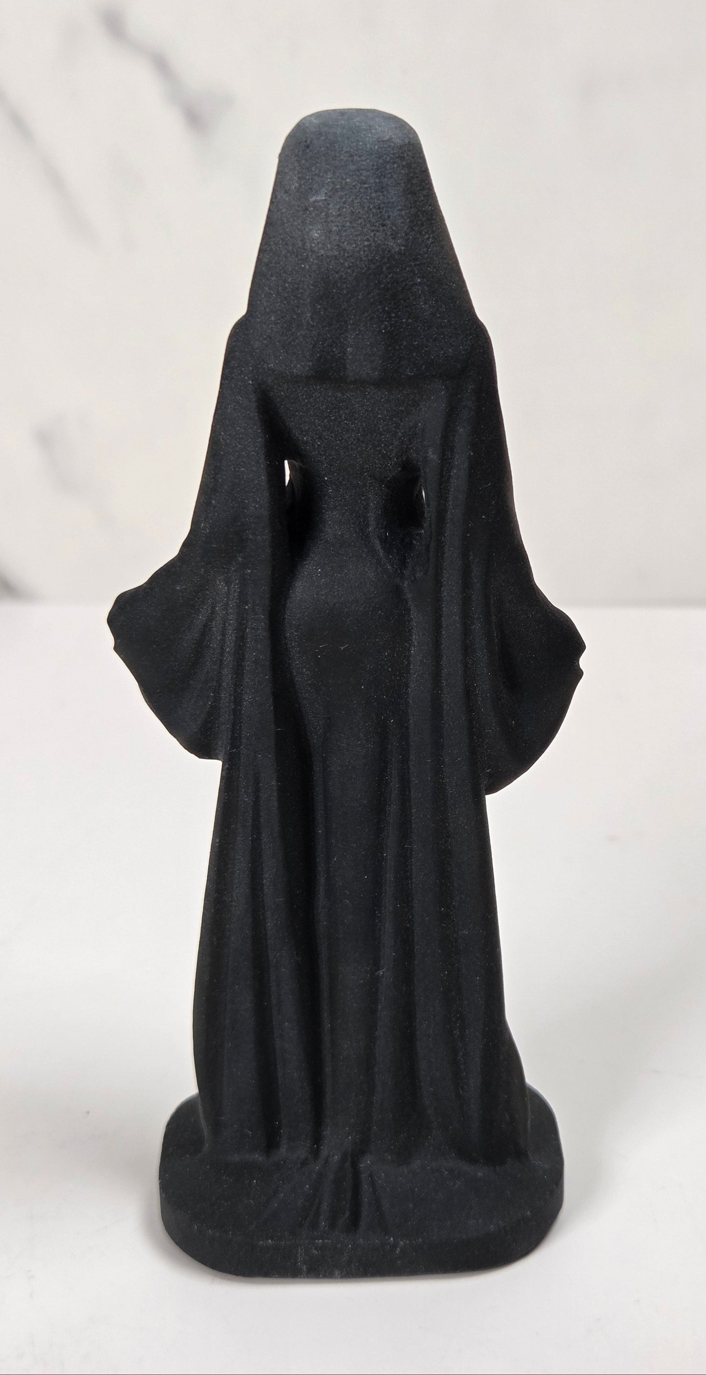 A small black figurine of a hooded robed figure, shown from the back, standing on a white surface against a light gray marble-patterned background.