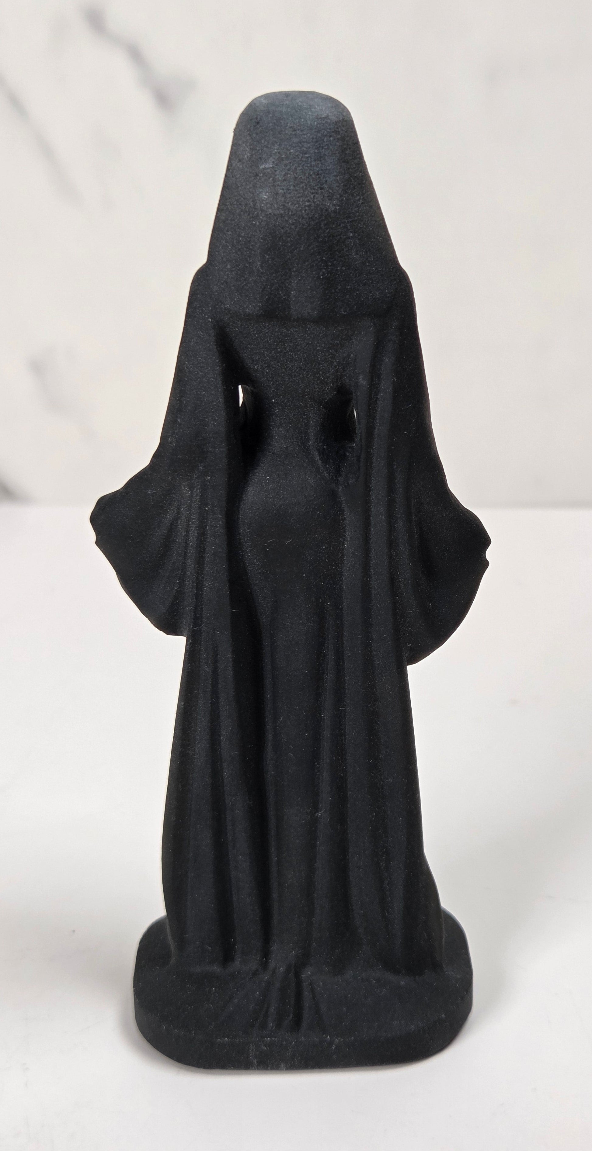 A small black figurine of a hooded robed figure, shown from the back, standing on a white surface against a light gray marble-patterned background.