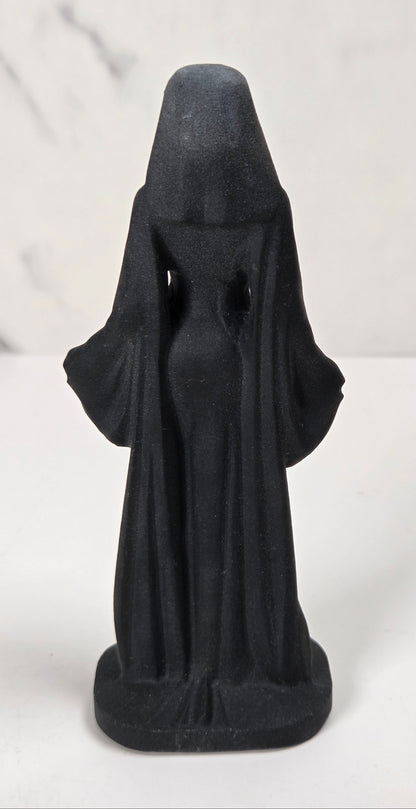 A small black figurine of a hooded robed figure, shown from the back, standing on a white surface against a light gray marble-patterned background.
