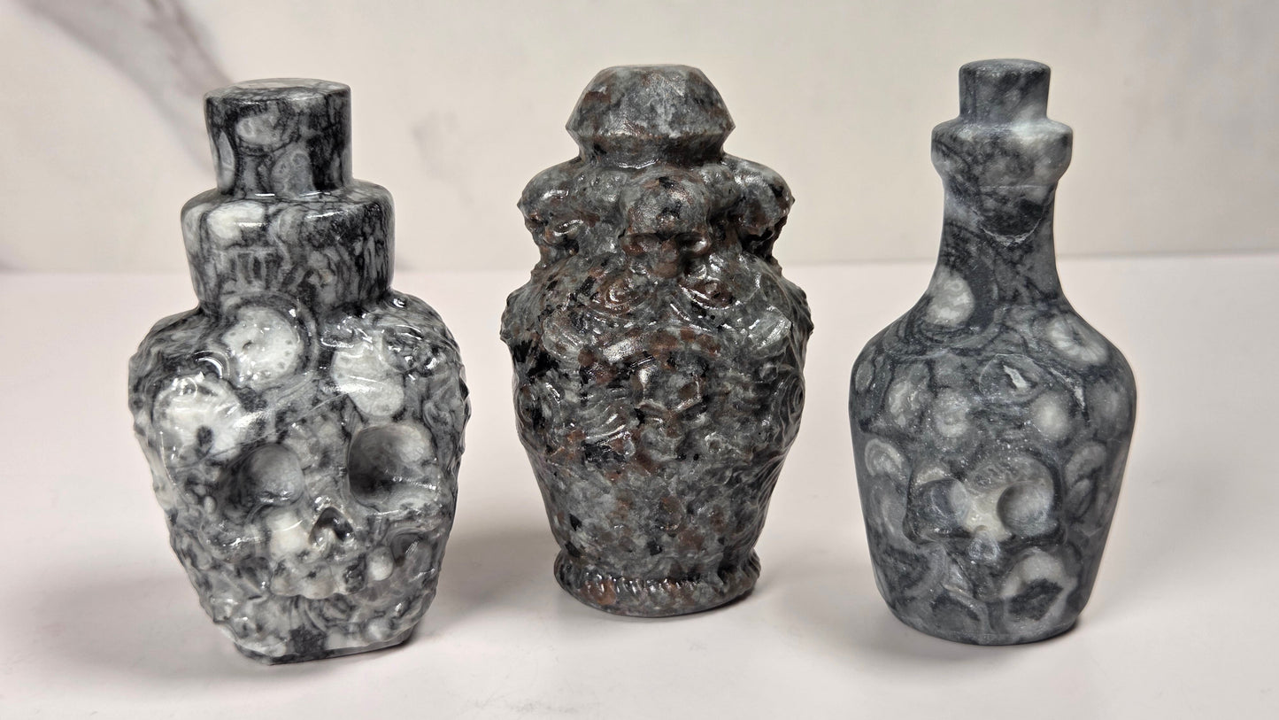 Three small decorative bottles or vases made of mottled stone or resin, shown standing upright on a white surface against a light neutral background. The left and right pieces are gray and white marbled, while the center piece is darker gray with some brownish undertones.