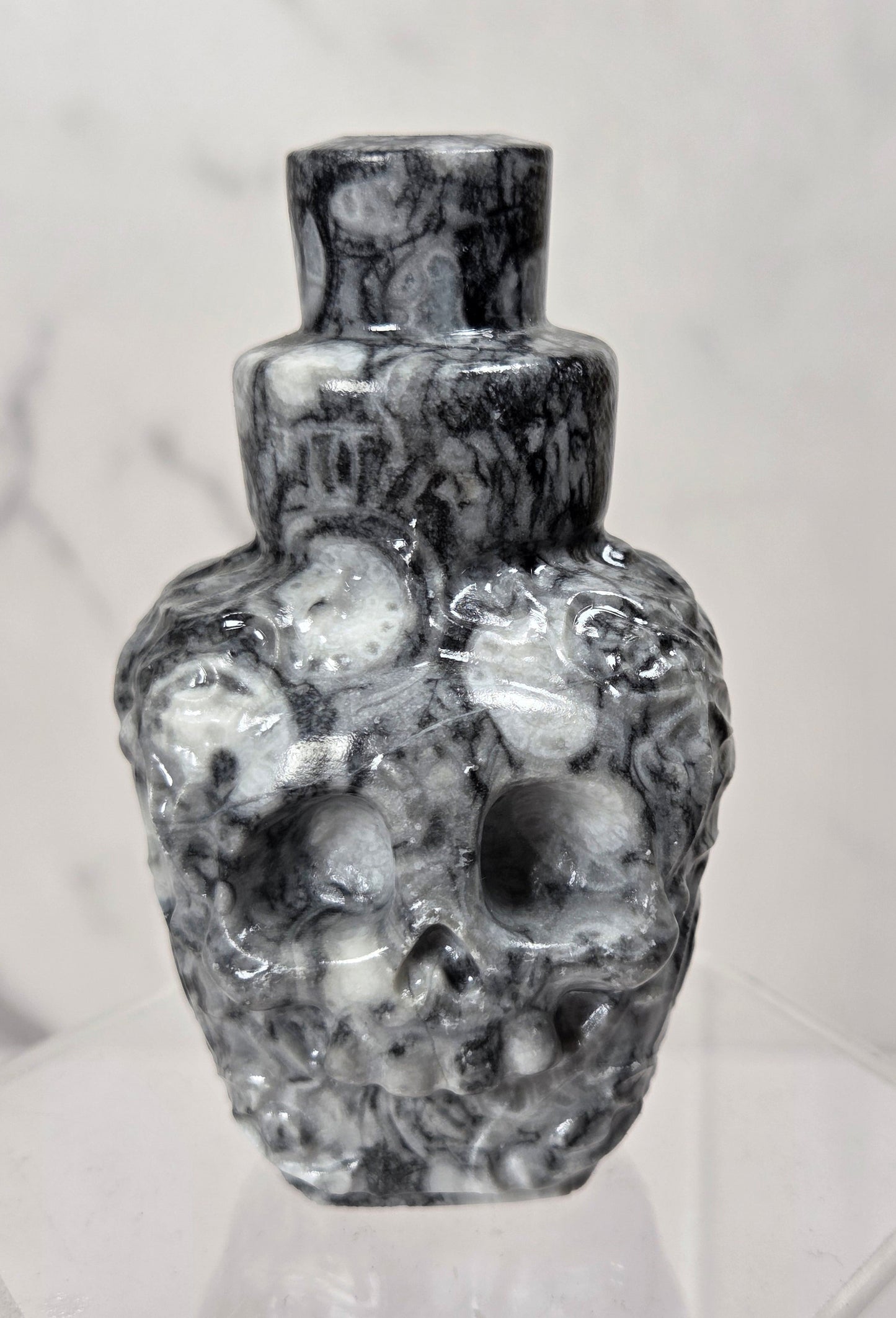 A small sculptural ceramic vase with a glossy black and white marbled pattern, featuring a textured, organic surface and a short neck, displayed on a clear acrylic stand against a light gray marble background.