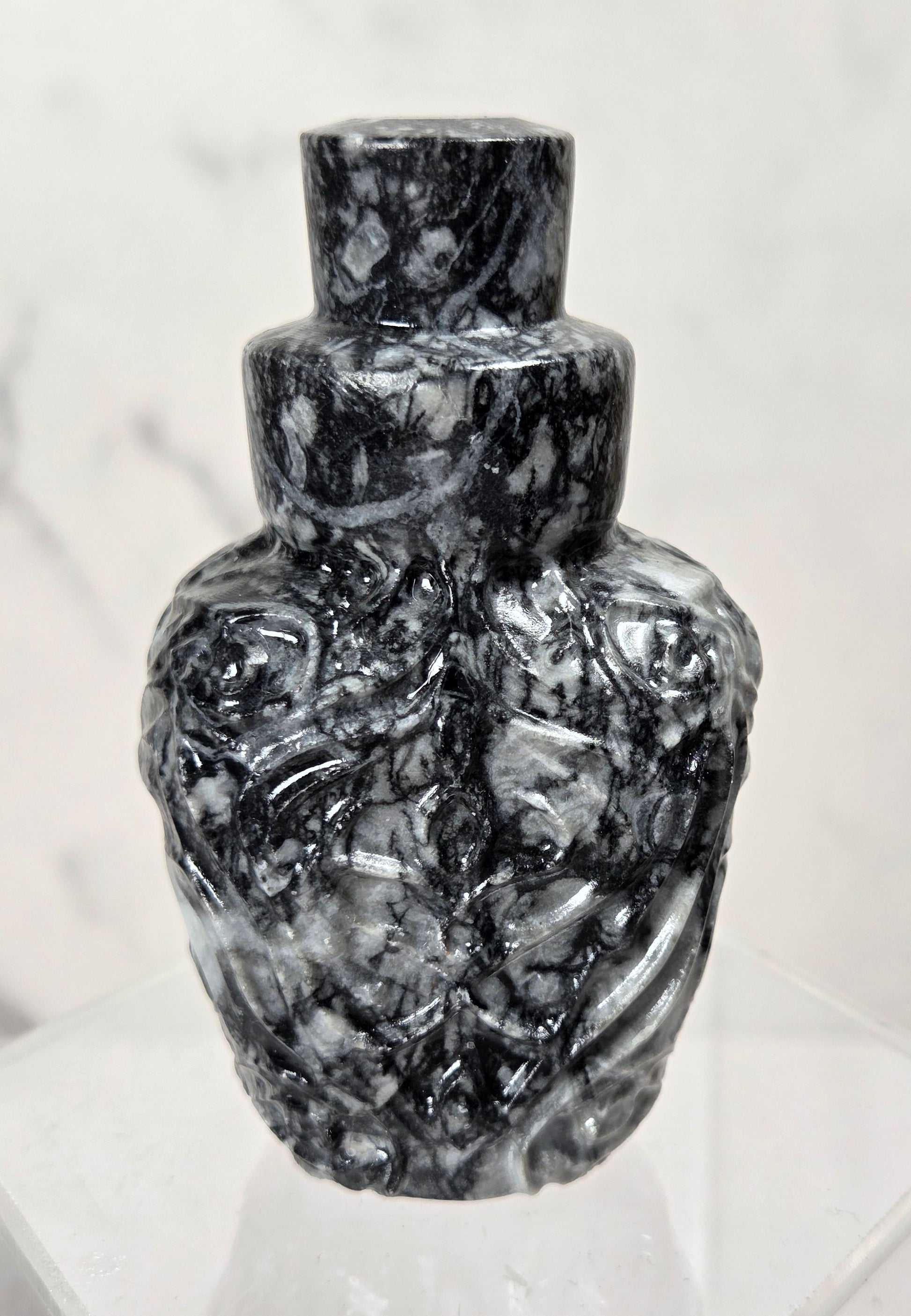 A small sculptural bottle or vase made of polished black and white marbled stone, displayed on a clear acrylic stand against a light marble-patterned background.