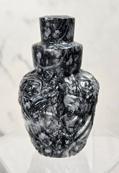 A small sculptural bottle or vase made of polished black and white marbled stone, displayed on a clear acrylic stand against a light marble-patterned background.