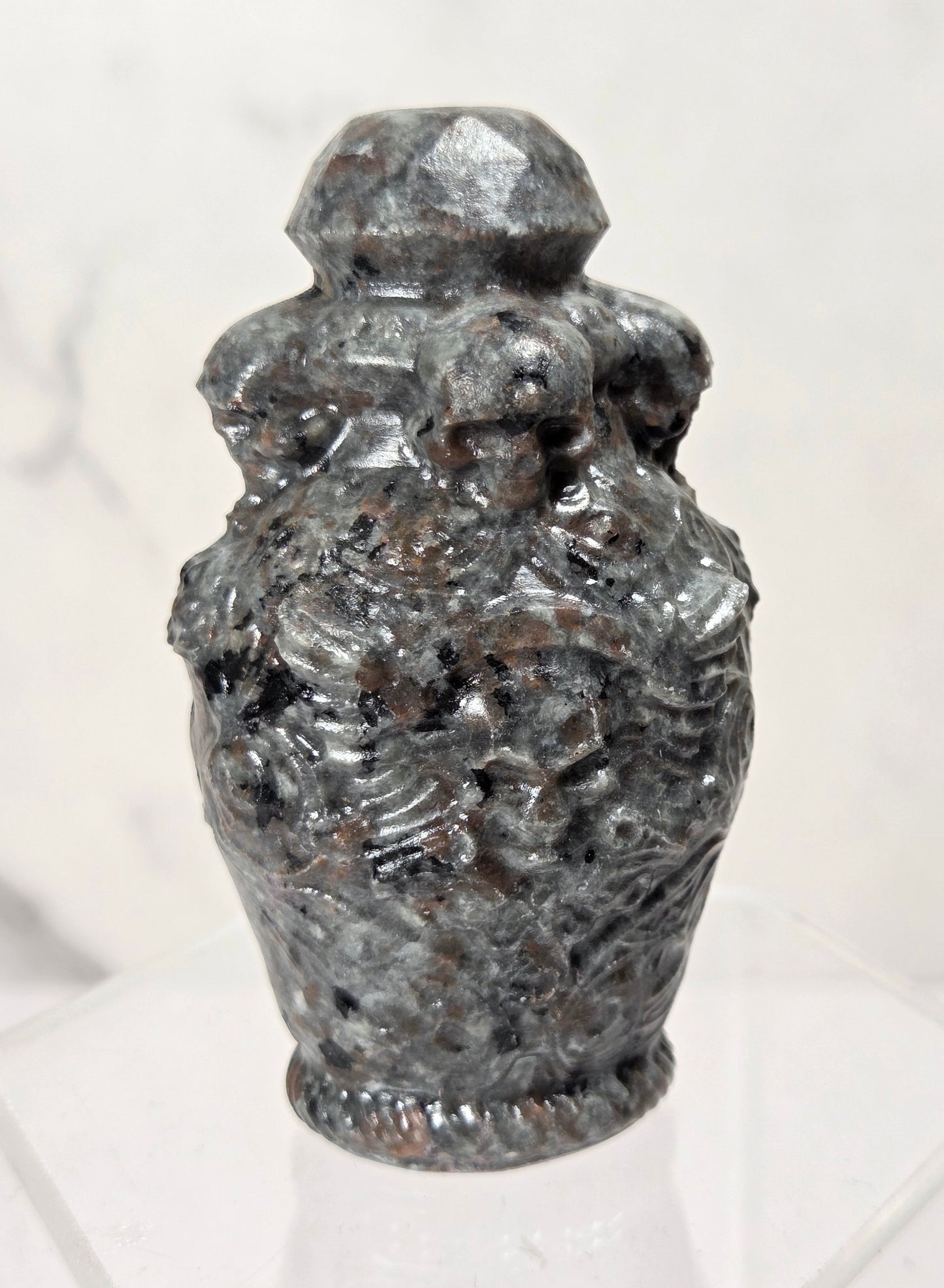 A small carved stone urn or vase with a faceted lid, displayed on a clear acrylic stand against a light marble-patterned background.