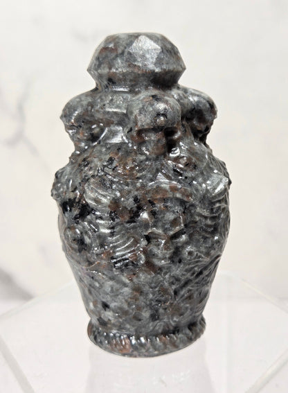 A small carved stone urn or vase with a faceted lid, displayed on a clear acrylic stand against a light marble-patterned background.