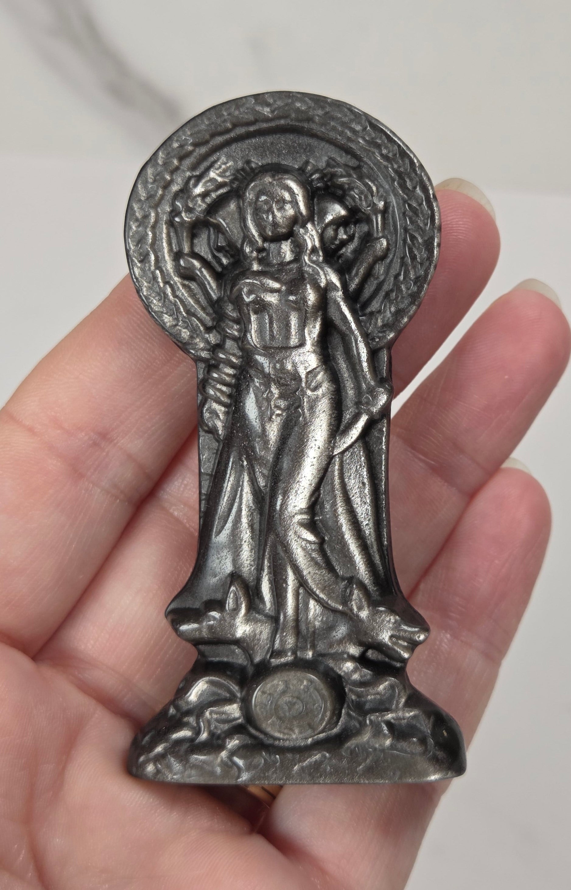 A hand holding a small silver-toned religious figurine of Hecate, shown against a light marble-patterned background.
