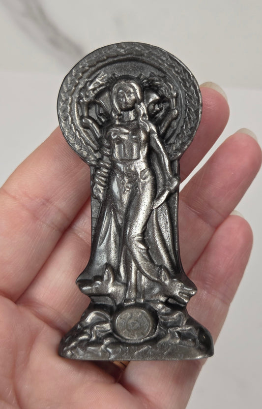 A hand holding a small silver-toned religious figurine of Hecate, shown against a light marble-patterned background.
