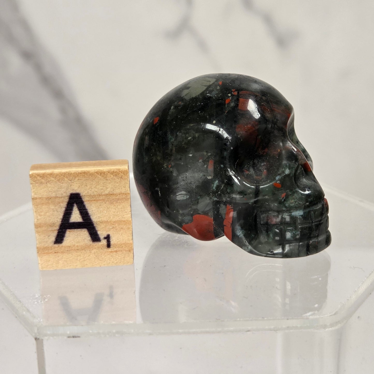 A polished dark  jasper skull sculpture displayed on a clear acrylic stand next to a wooden Scrabble tile showing the letter A with a subscript 1, set against a light gray marble-patterned background.