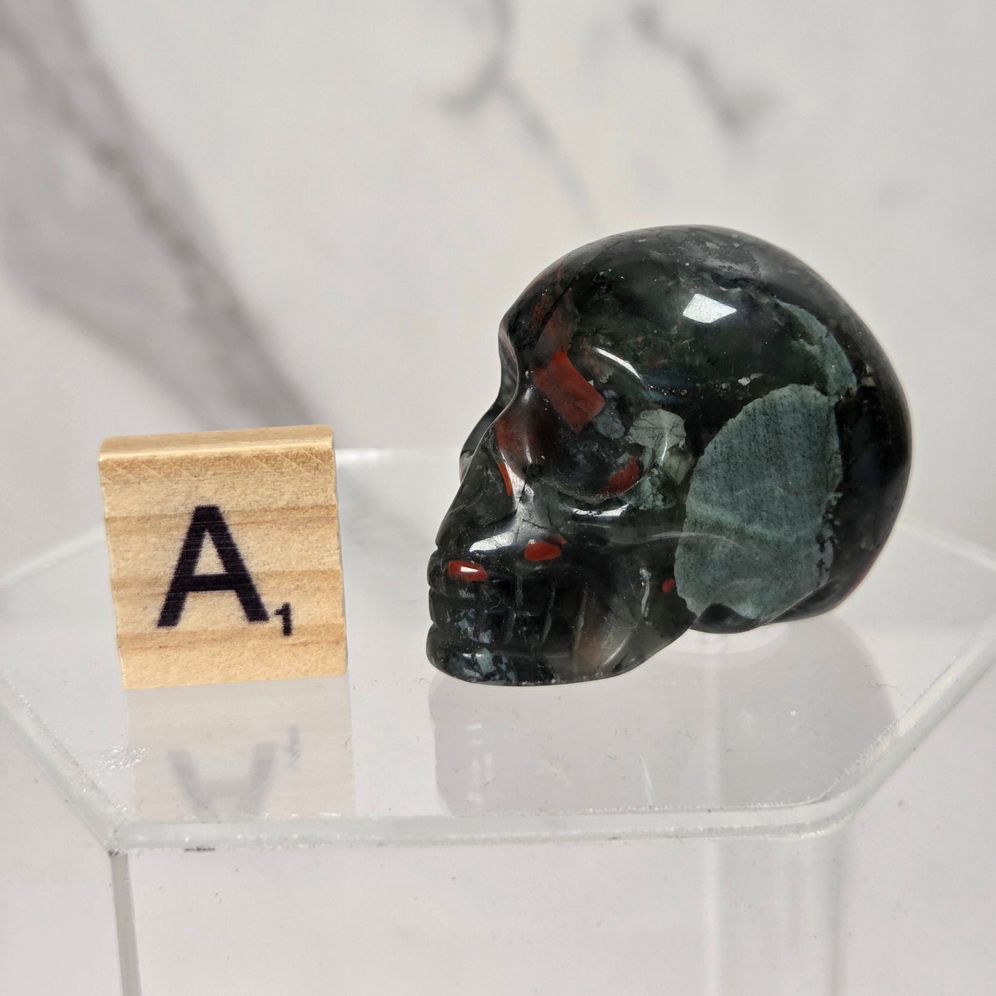 A polished black skull-shaped object with green and red inclusions, displayed on a clear acrylic stand next to a wooden Scrabble tile showing the letter A.