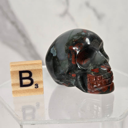 A polished carved skull made of dark marbled stone with black, gray, and reddish-brown veining, displayed on a clear acrylic stand next to a wooden Scrabble tile showing the letter B3.
