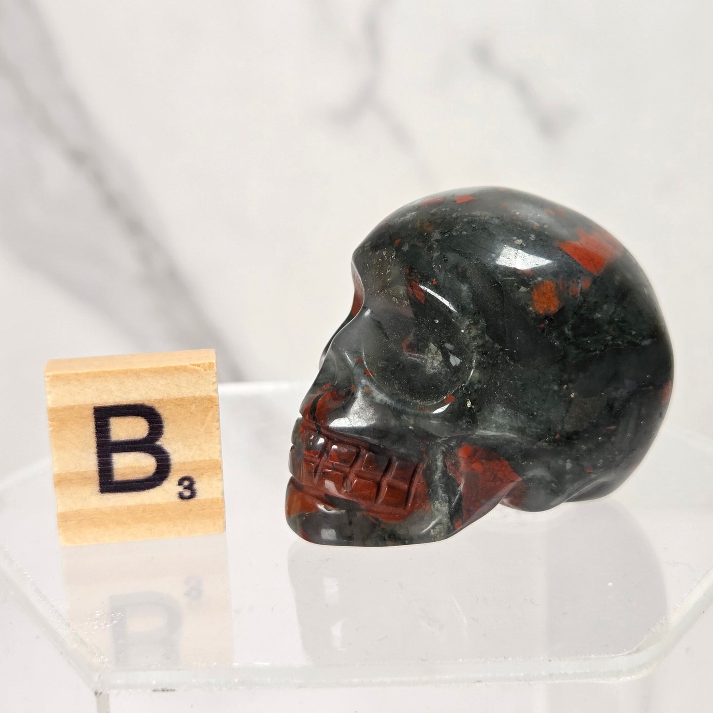 A polished black obsidian skull with red inclusions, displayed on a clear acrylic stand next to a wooden Scrabble tile showing the letter B3, against a white marble background.