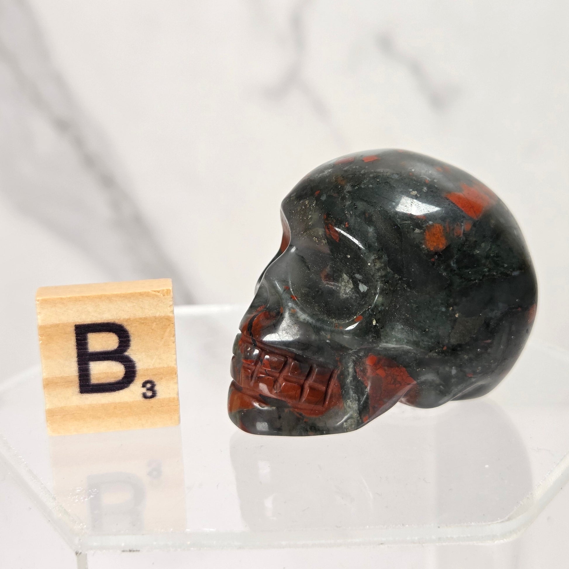 A polished black obsidian skull with red inclusions, displayed on a clear acrylic stand next to a wooden Scrabble tile showing the letter B3, against a white marble background.