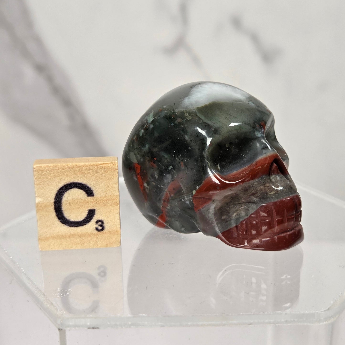 A polished carbonite skull figurine with dark green, black, and red banding, displayed on a clear acrylic stand next to a Scrabble tile showing 'C3' on a light marble background.