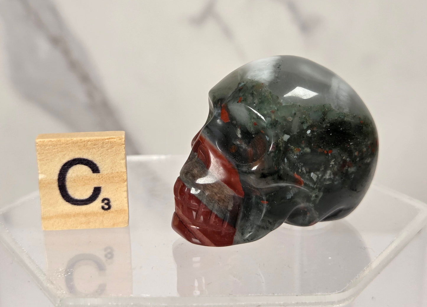 A polished red jasper skull figurine with dark green and black inclusions, displayed on a clear acrylic stand next to a Scrabble tile showing the letter C with a 3.