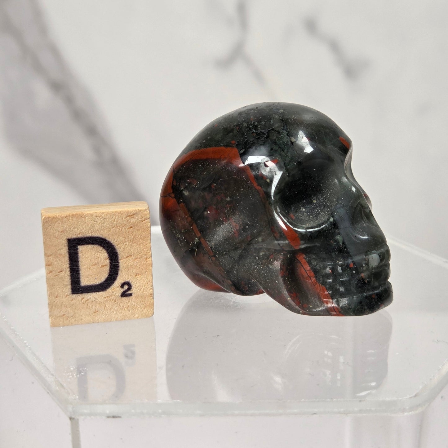 A polished black skull-shaped crystal or stone ornament with red and white marbling, displayed on a clear acrylic stand next to a small Scrabble tile showing D².