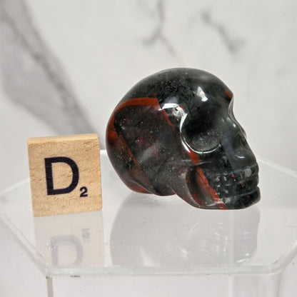 A polished black skull-shaped crystal or stone ornament with red and white marbling, displayed on a clear acrylic stand next to a small Scrabble tile showing D².