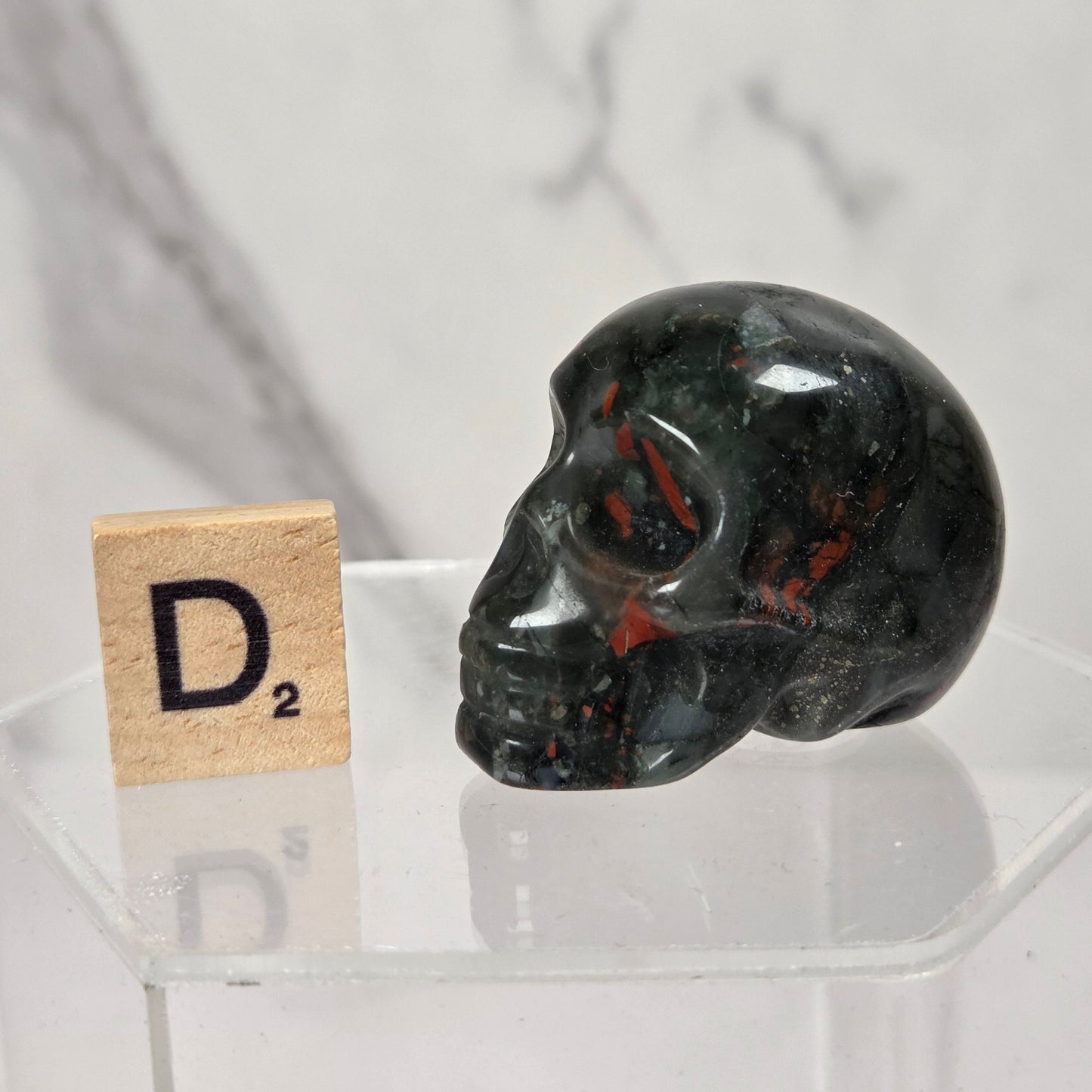 A polished dark green and black jasper skull with red inclusions, displayed on a clear acrylic stand next to a wooden Scrabble tile showing the letter D2, against a light gray marble background.