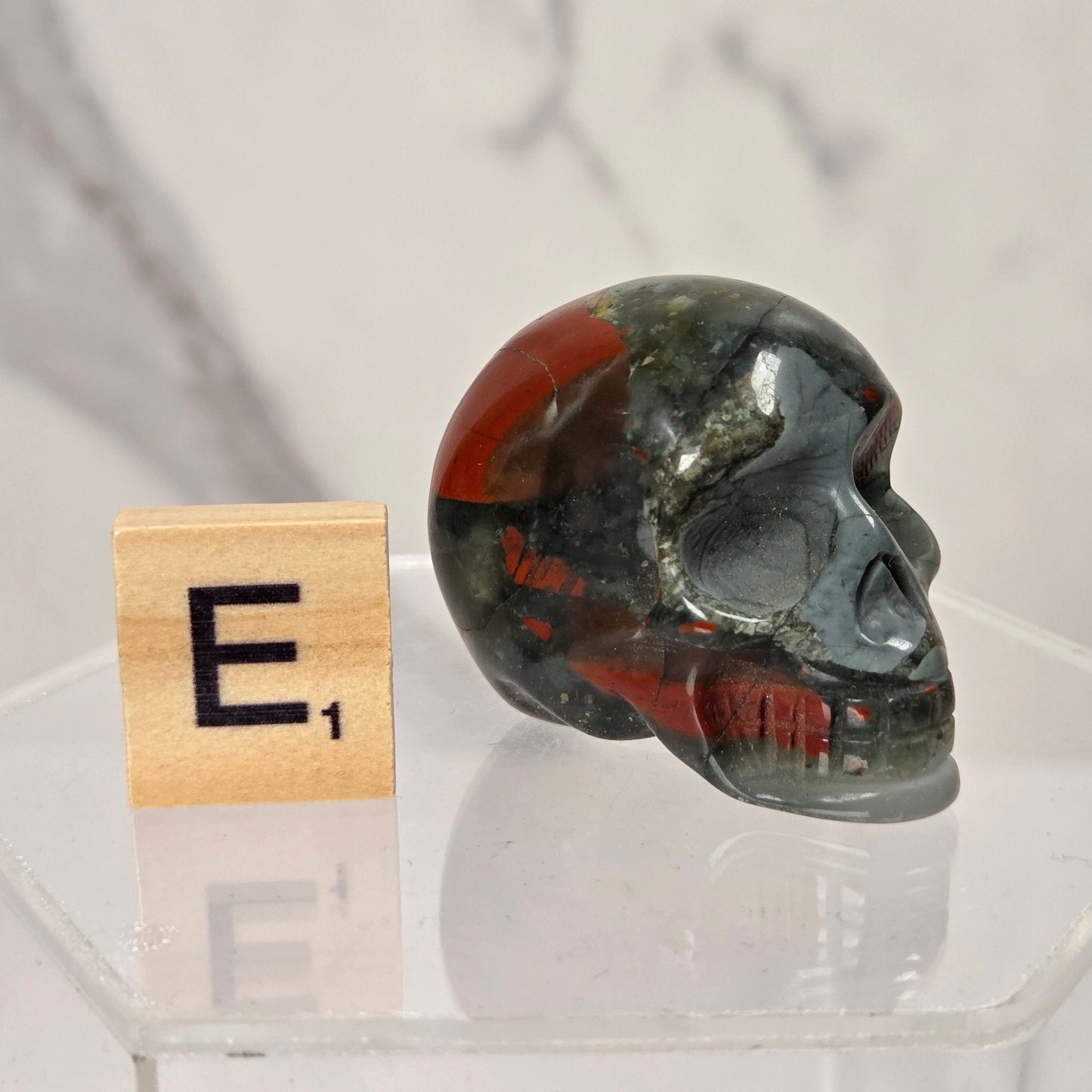 A polished red jasper skull sits on a clear acrylic display base next to a wooden Scrabble tile showing the letter 'E' and value of 1, against a light marble-patterned background.