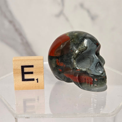 A polished red jasper skull sits on a clear acrylic display base next to a wooden Scrabble tile showing the letter 'E' and value of 1, against a light marble-patterned background.