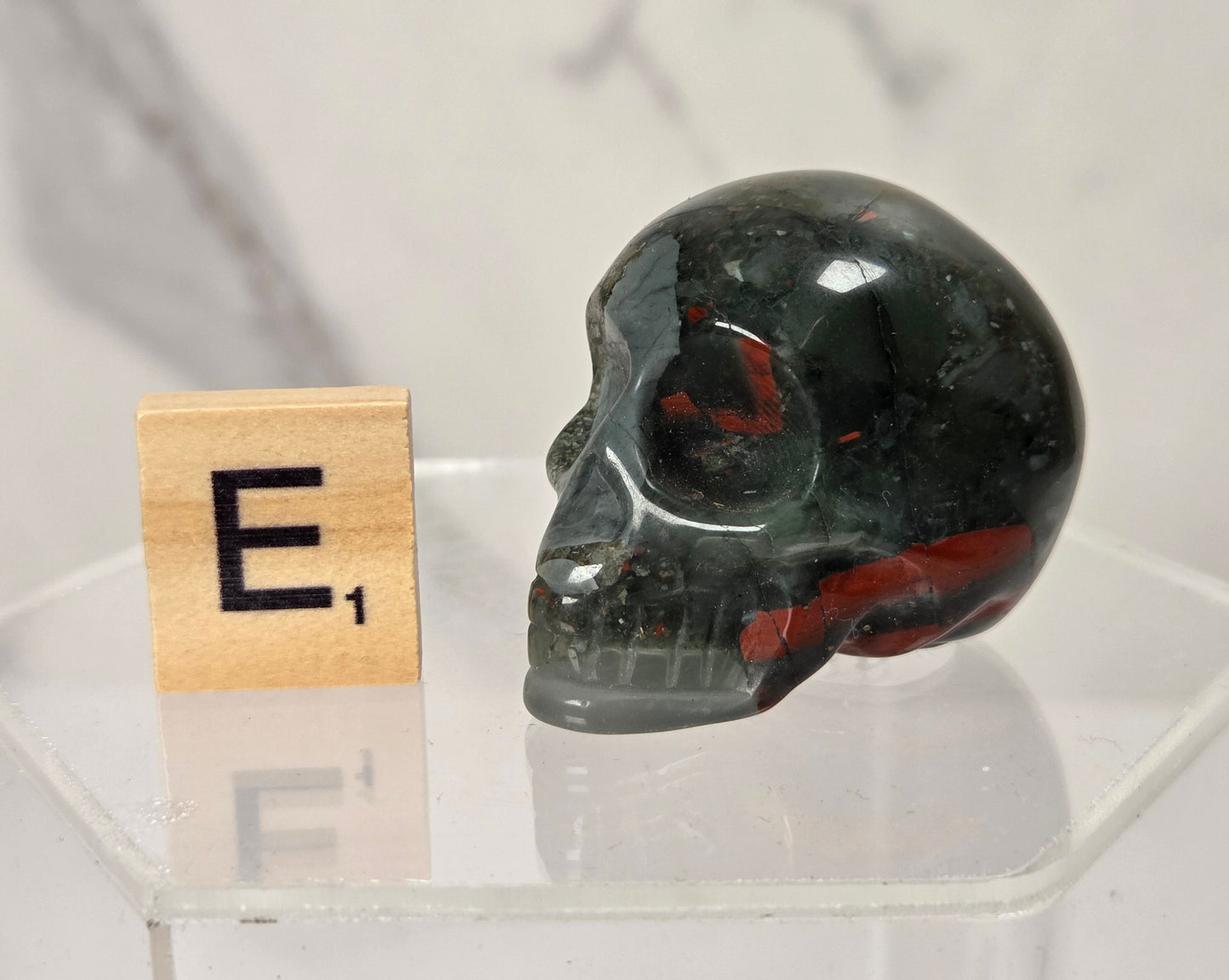 A polished dark green skull with red inclusions, displayed on a clear acrylic stand next to a wooden Scrabble tile showing the letter 'E'.