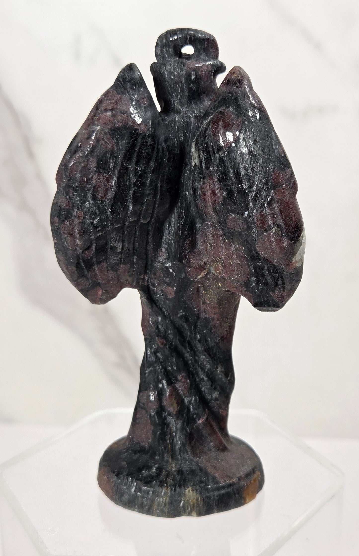 A small dark stone angel figurine with outstretched wings and a halo, displayed on a clear acrylic stand against a light marble-patterned background.