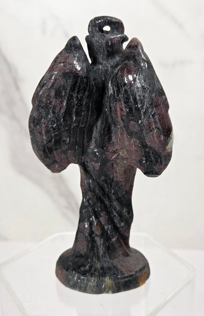 A small dark stone angel figurine with outstretched wings and a halo, displayed on a clear acrylic stand against a light marble-patterned background.