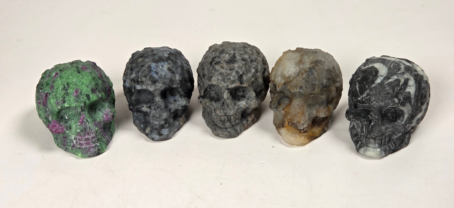 Five small carved skull-shaped objects arranged in a row on a light surface. The skulls have varied natural stone-like colors and marbled patterns, including green, blue-gray, dark gray, tan-and-brown, and black-and-white marbling.