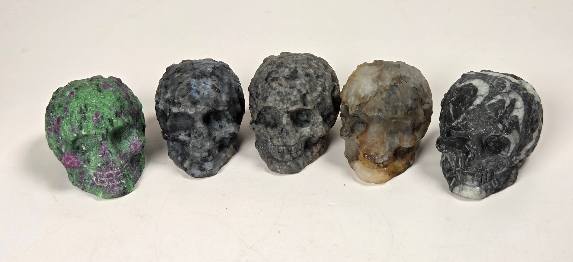 Five small carved skull-shaped objects arranged in a row on a light surface. The skulls have varied natural stone-like colors and marbled patterns, including green, blue-gray, dark gray, tan-and-brown, and black-and-white marbling.