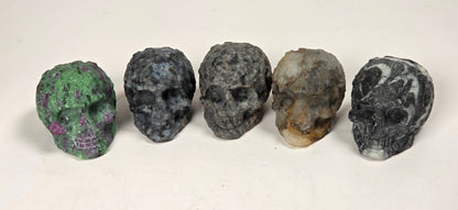 Five small carved skull-shaped objects arranged in a row on a light surface. The skulls have varied natural stone-like colors and marbled patterns, including green, blue-gray, dark gray, tan-and-brown, and black-and-white marbling.