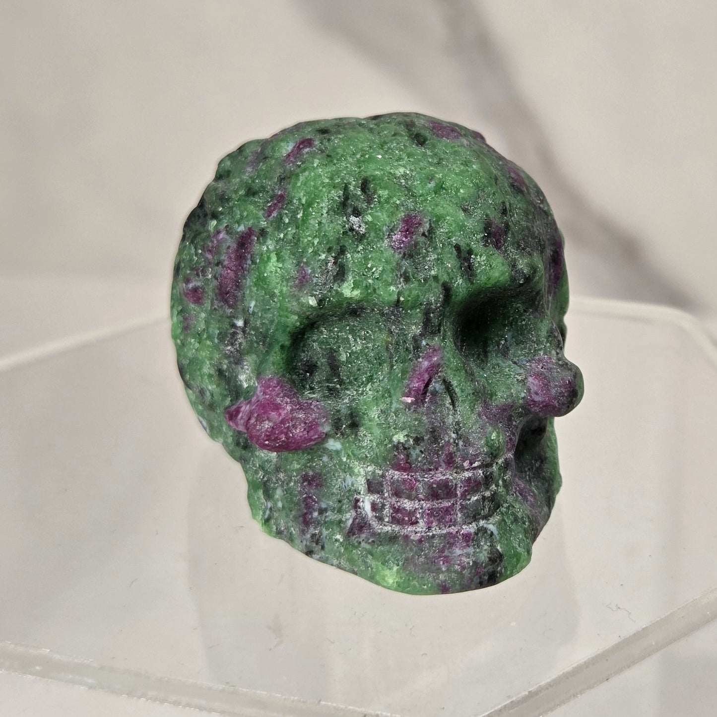 A small carved skull made from green and purple marbled stone, displayed on a clear acrylic stand against a light gray marble-patterned background.