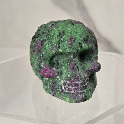 A small carved skull made from green and purple marbled stone, displayed on a clear acrylic stand against a light gray marble-patterned background.