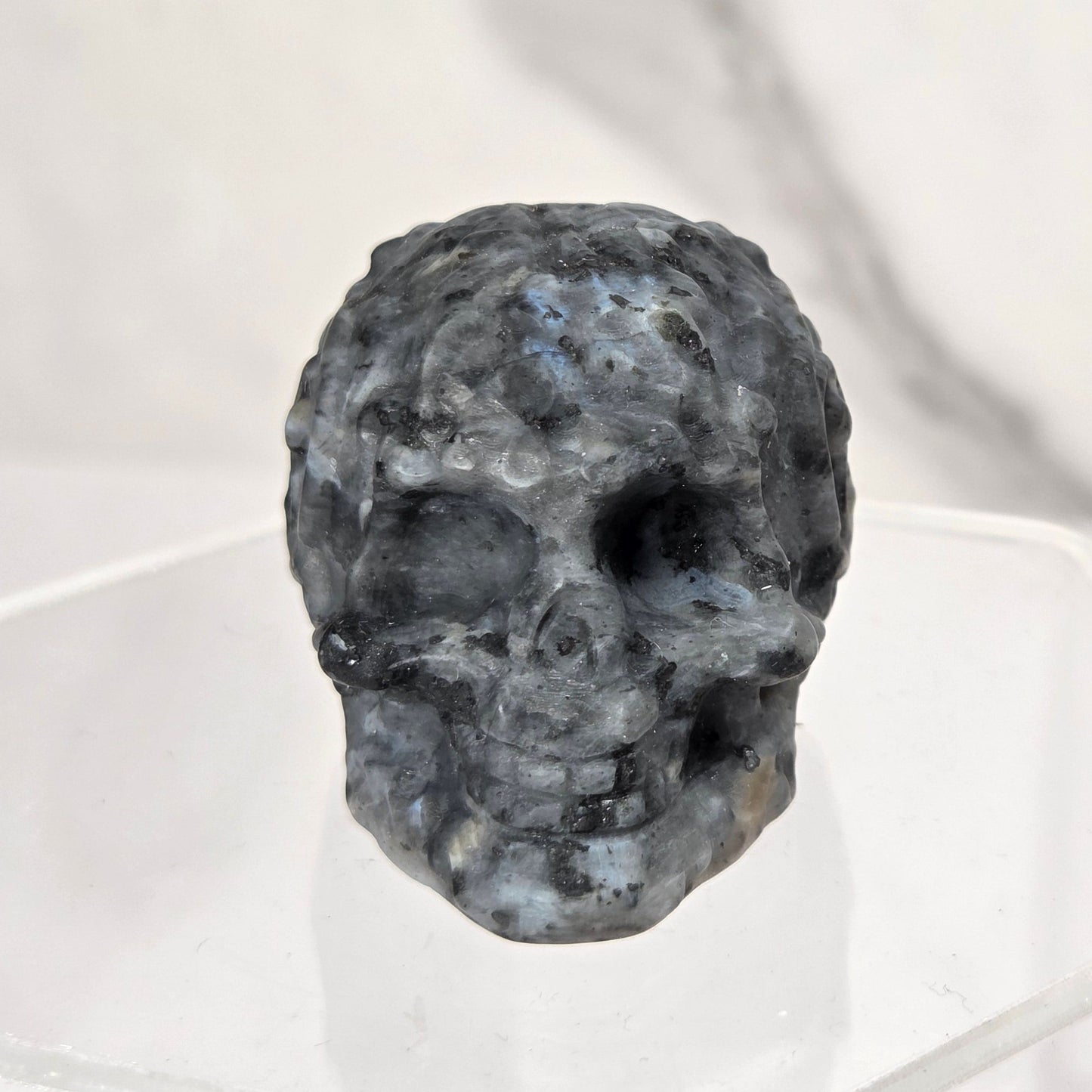 A small carved skull made of speckled dark stone, displayed on a clear acrylic stand against a light marble-patterned background.