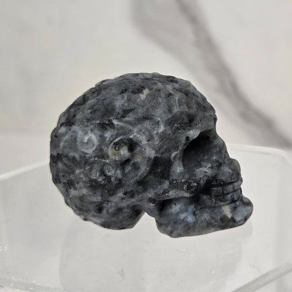 A small dark gray skull-shaped object displayed on a clear acrylic stand against a light marble-patterned background.