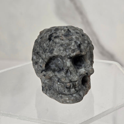 A small gray stone skull figurine displayed on a clear acrylic stand against a light marble-patterned background.