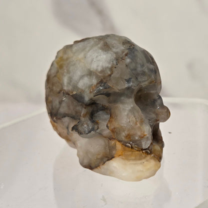 A small polished stone or crystal skull displayed on a clear acrylic stand against a soft light gray marble-like background.