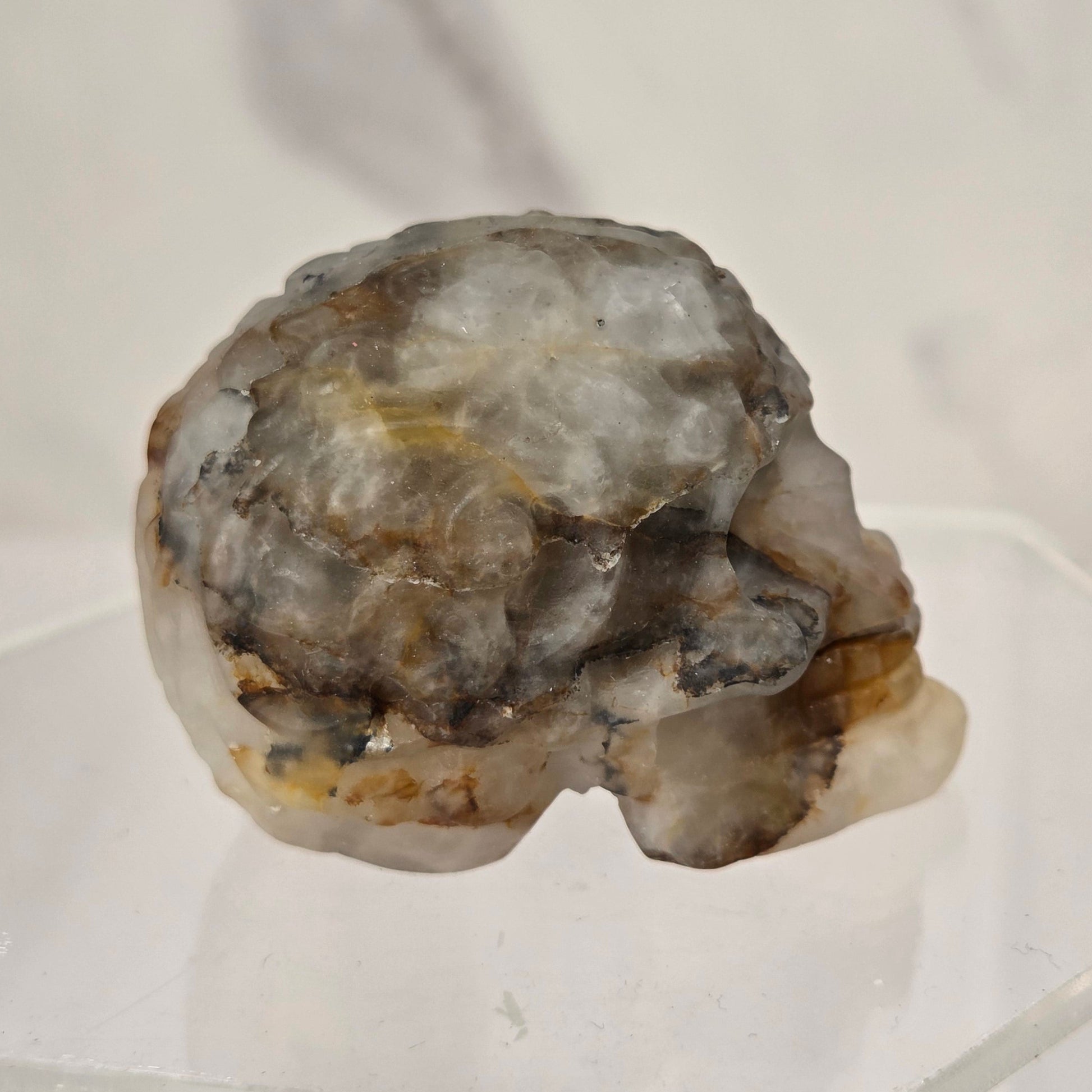 A raw, rough gemstone skull specimen with natural gray, white, and brown mineral textures, displayed on a clear acrylic stand against a light marble-patterned background.