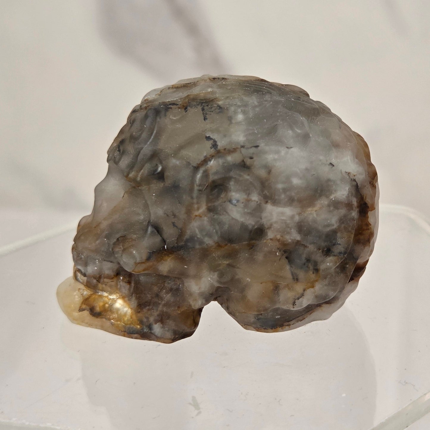 A raw, rough mineral specimen with a mix of gray, brown, and beige tones, displayed on a clear acrylic stand against a light gray marble-patterned background.