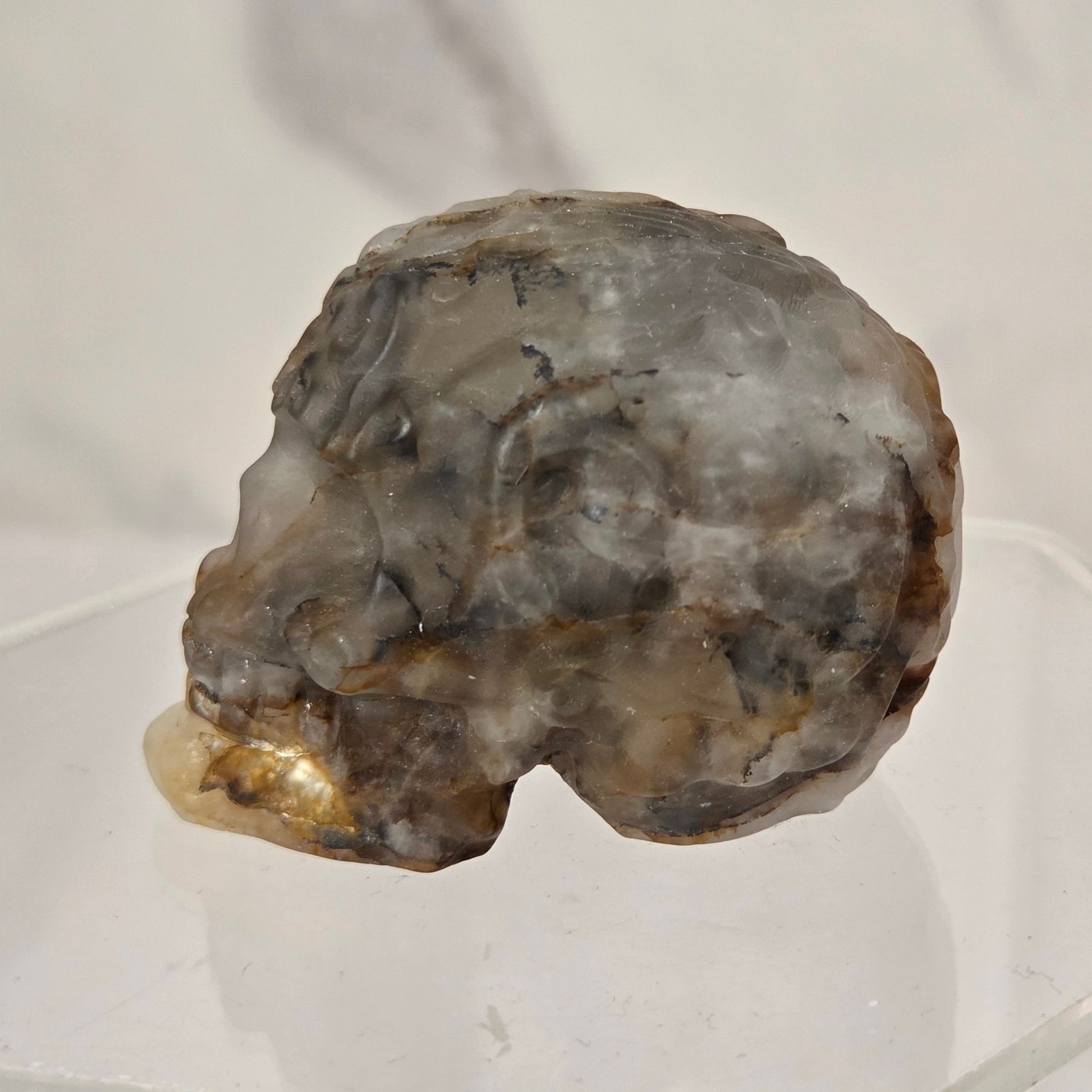 A raw, rough mineral specimen with a mix of gray, brown, and beige tones, displayed on a clear acrylic stand against a light gray marble-patterned background.