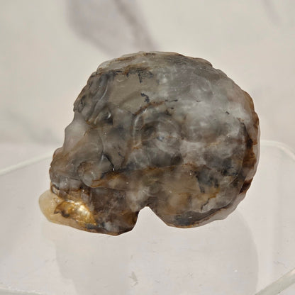 A raw, rough mineral specimen with a mix of gray, brown, and beige tones, displayed on a clear acrylic stand against a light gray marble-patterned background.