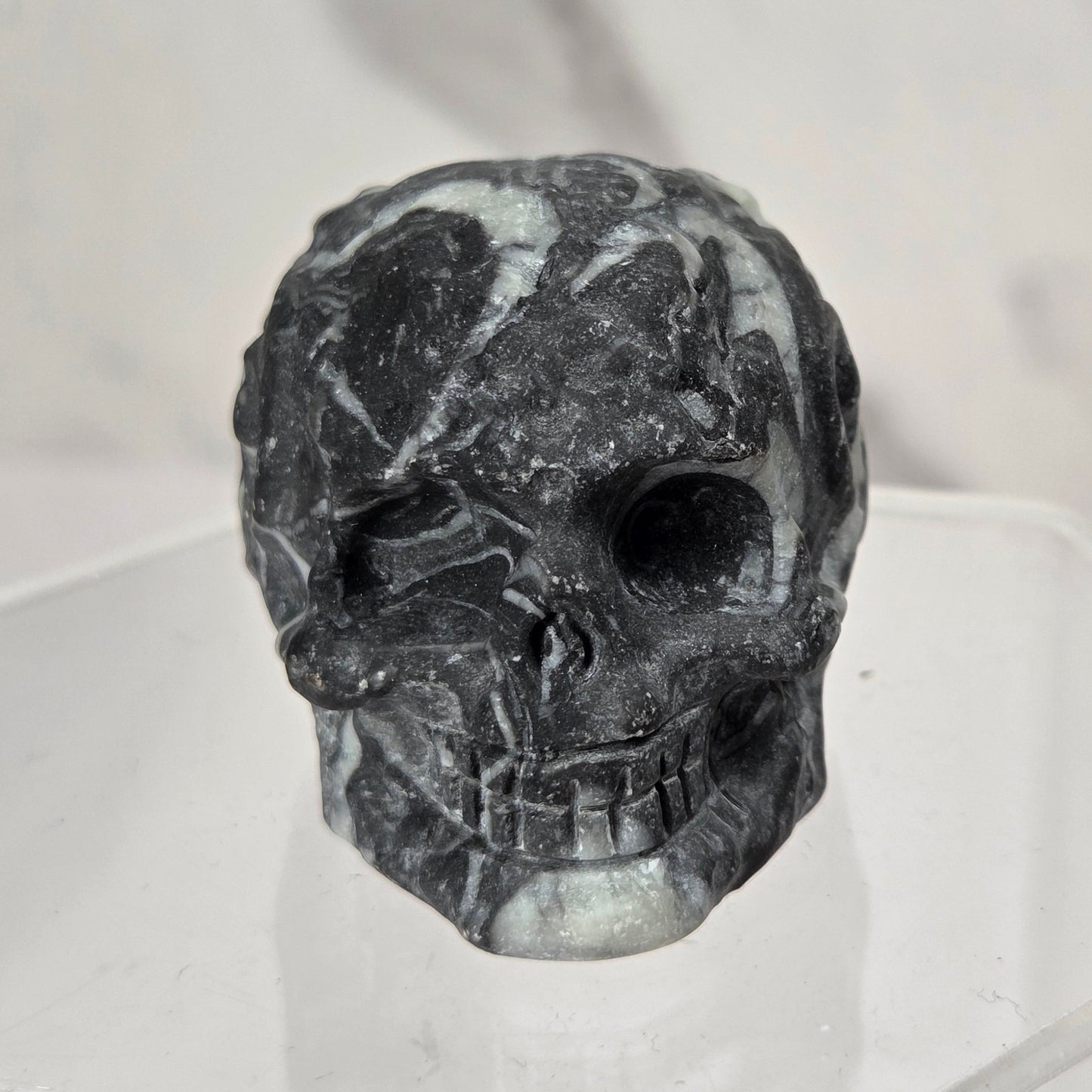 A carved black skull figurine with natural gray and white marbled patterns, displayed on a clear acrylic stand against a light gray marble-patterned background.